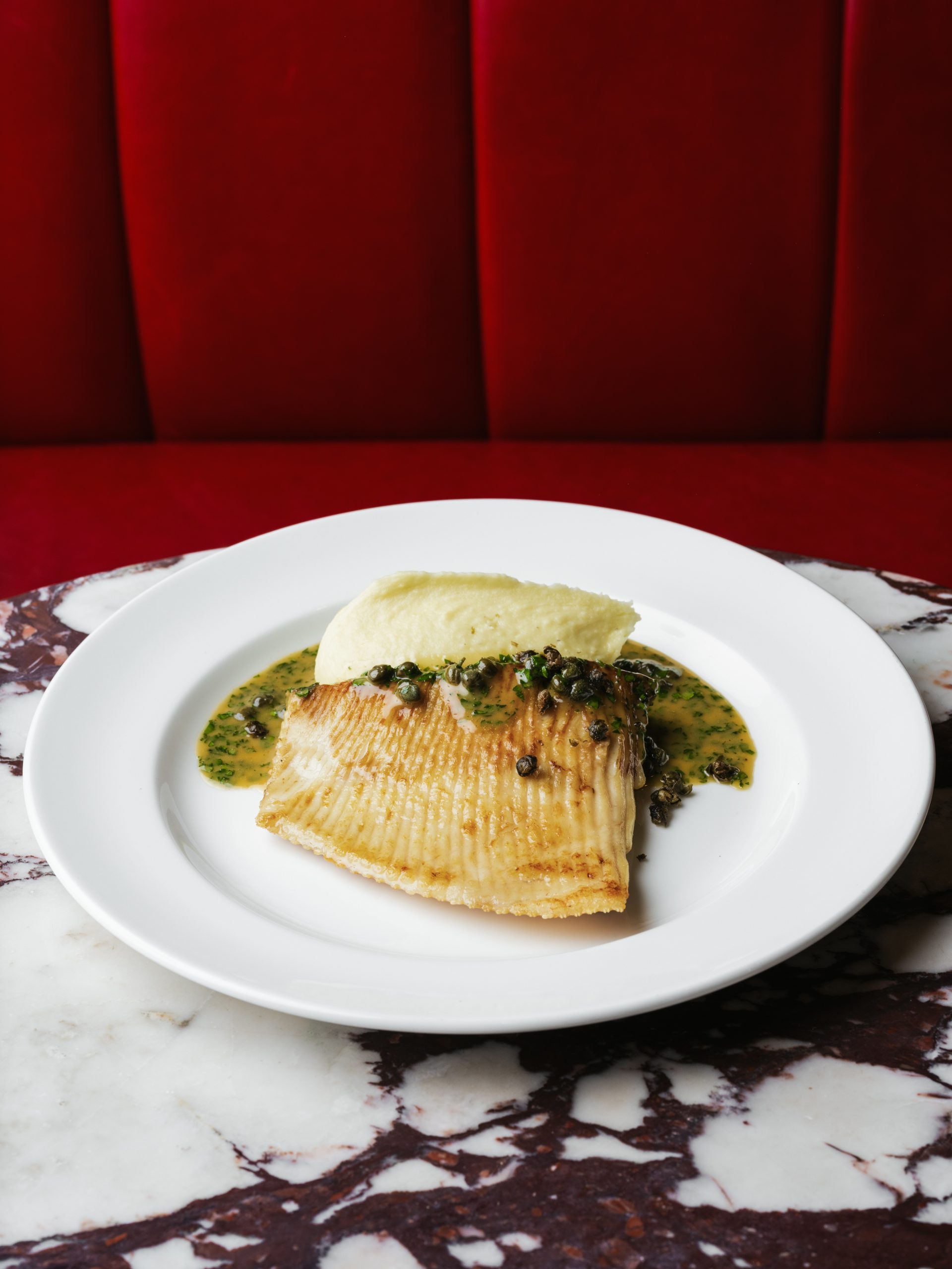Pan-fried Cornish skate with lemon butter, capers, parsley and potato purée on white plate against red leather banquette at Bistrot at Wild Honey London