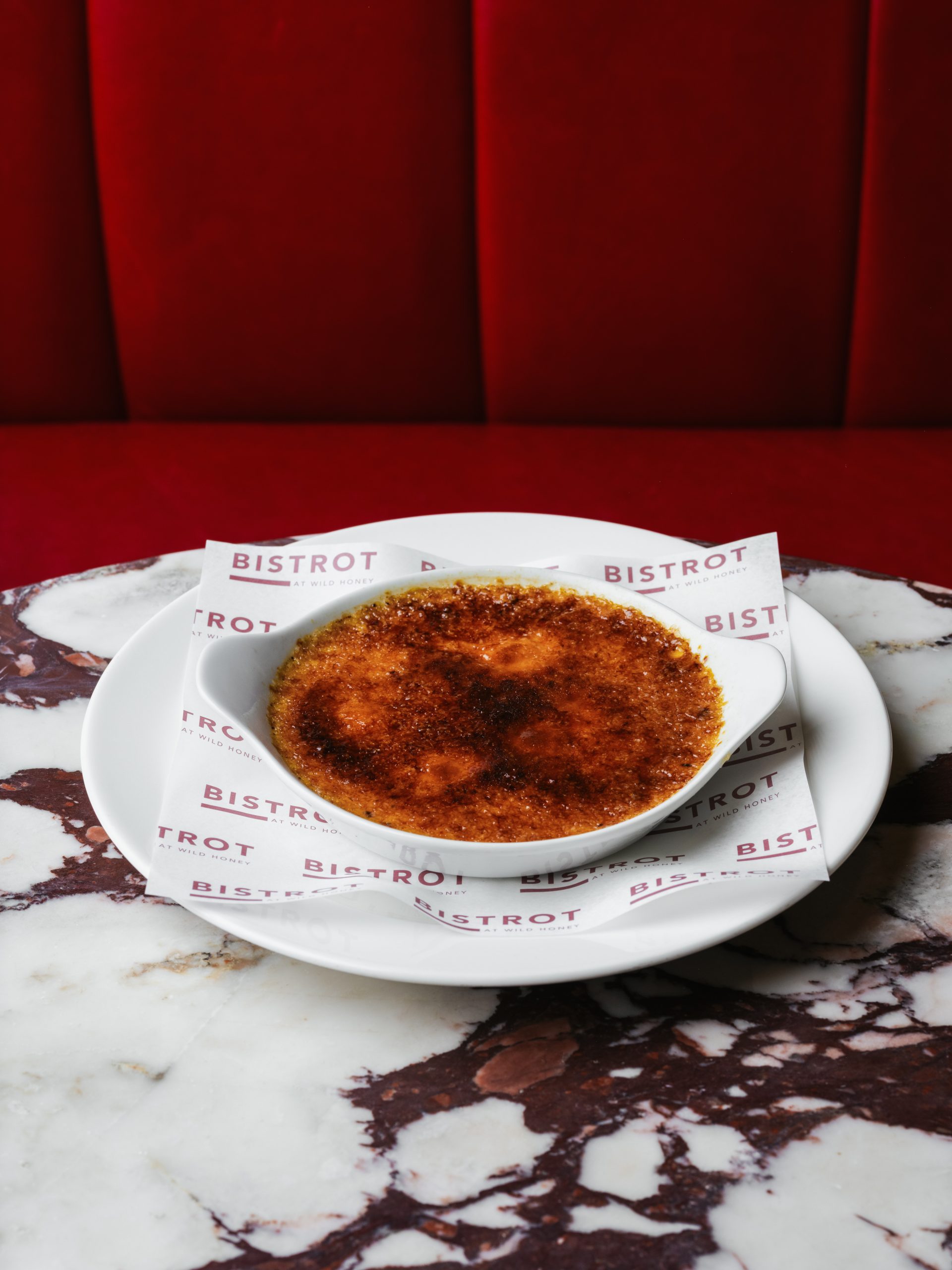 Crème brûlée in white ramekin with caramelised sugar crust on Bistrot at Wild Honey branded coasters against red leather banquette, marble table
