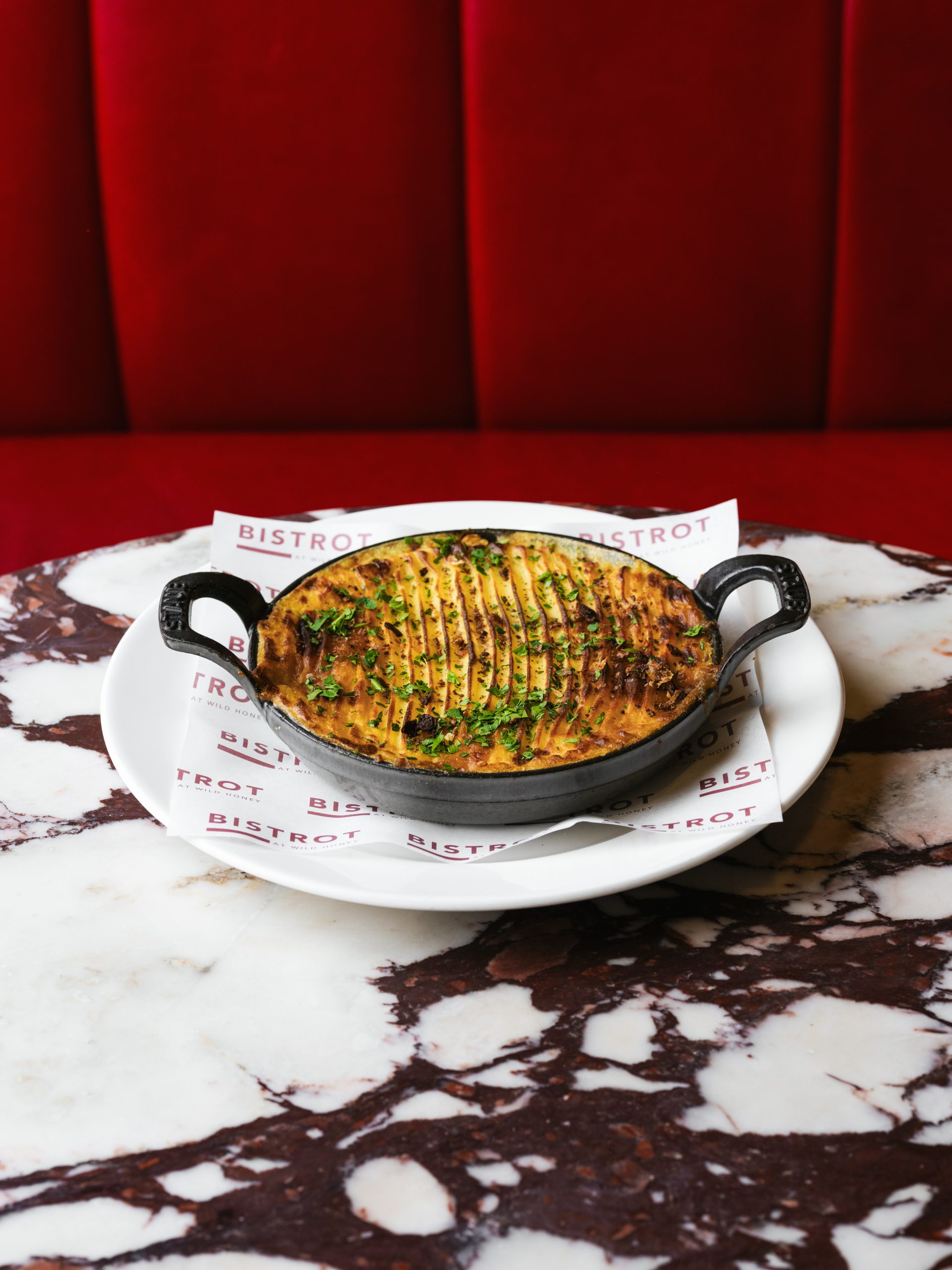 Shepherd's pie served in a black Staub cast-iron dish with piped mashed potato top, herb garnish and Bistrot at Wild Honey branded paper on marble table