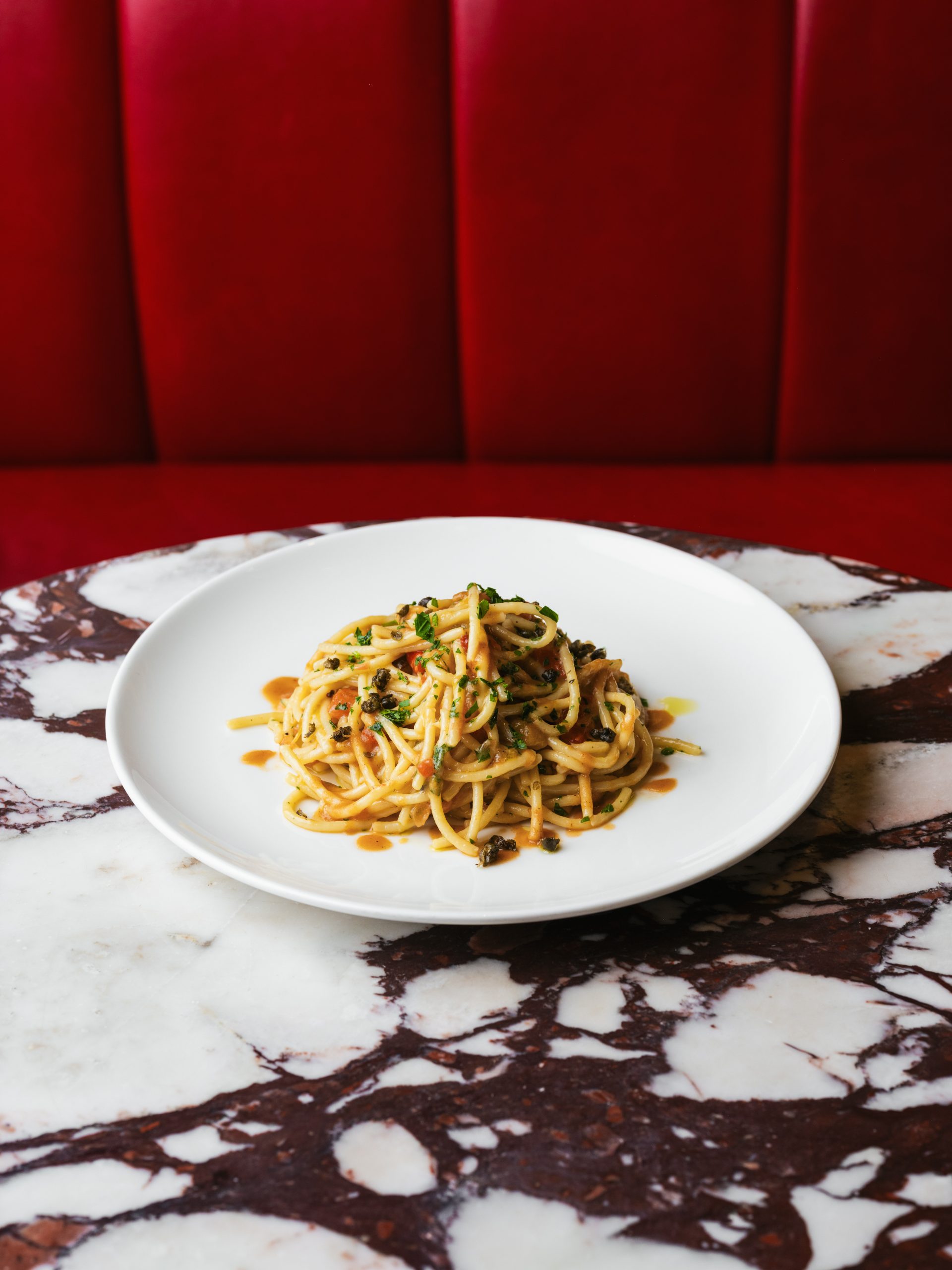 Spaghetti with San Marzano tomatoes, sweet peppers and capers on white plate against red leather banquette on marble table at Bistrot at Wild Honey London